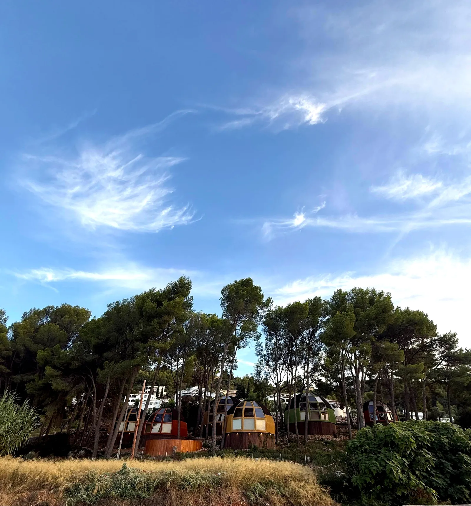 Las seis casas domo geodésicas de AUMARA en colores naranja, amarillo, verde y rojo bajo un amplio cielo azul en Benidoleig, Alicante (Costa Blanca)