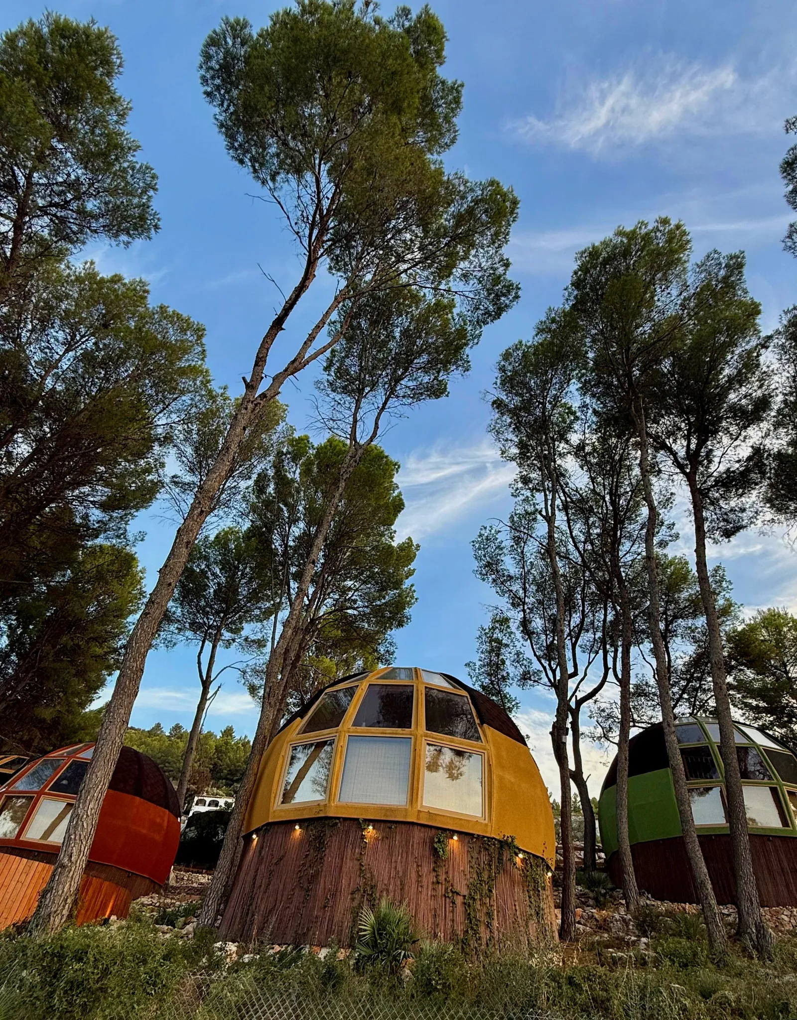 Casa domo geodésica amarilla de AUMARA con grandes ventanales y base de madera iluminada, junto a domos naranja y verde, en Benidoleig, Alicante (Costa Blanca)