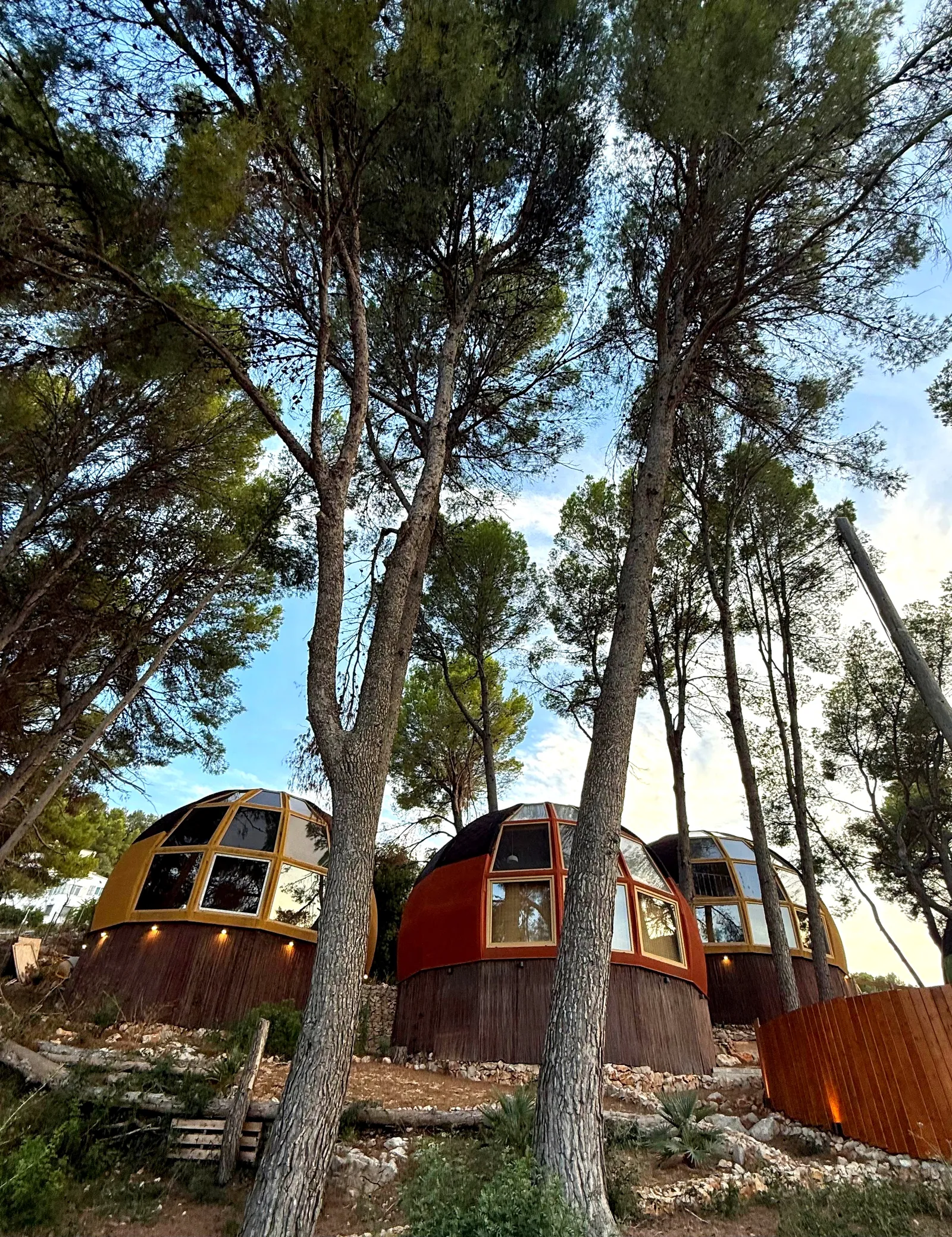Tres casas domo geodésicas de AUMARA, amarilla, naranja y ocre, con bases de madera y luces cálidas al atardecer en Benidoleig, Alicante (Costa Blanca)